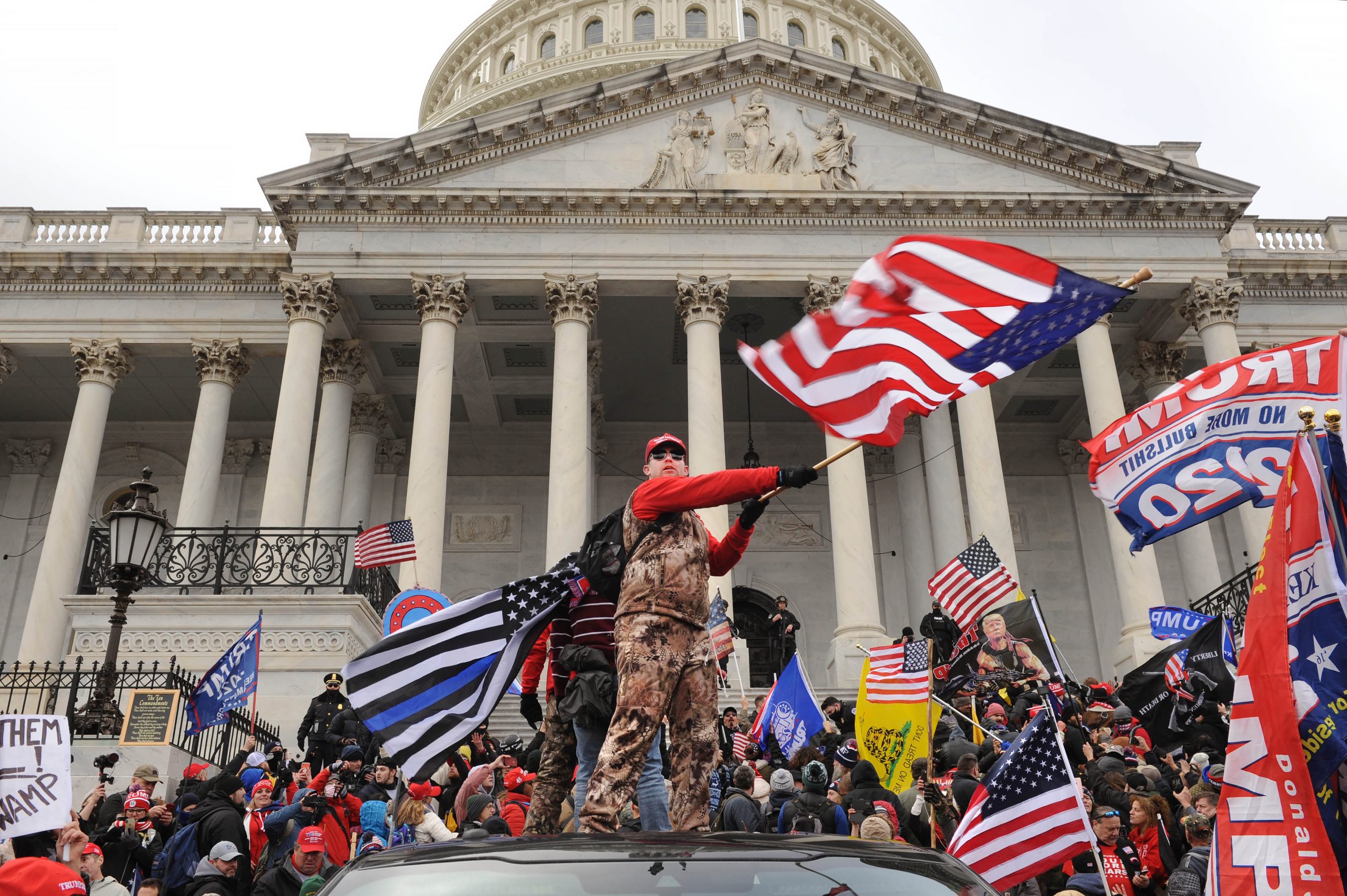 Overall shot of demonstrators on U.S. Capitol steps, man standing on top of a car waving flag, policeman in background at top, second policeman at left. Note QAnon (large Q) symbol and Blue Lives Matter flag (blue, white and black).