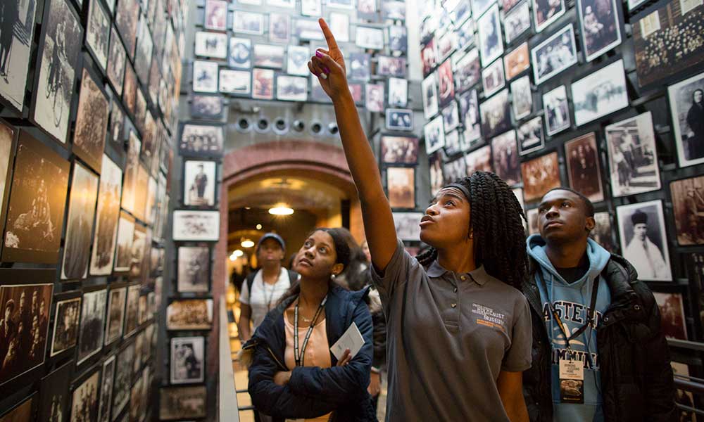 A guide leads a group of teenagers through a room in the Holocaust Museum whose walls are completely covered in historical photographs, pointing to a photo and telling the teenagers about it. Holocaust Education in action.