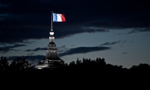 The French flag, made up of red, white and blue stripes, sits atop a tall buidling. Most of the flag's surroundings are in the shadows of the night. A light shines only on the flag.