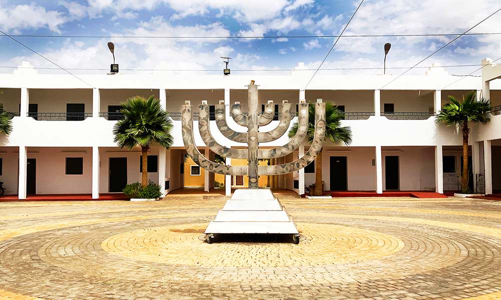 The photograph is visually divided into even horizonatal thirds. The top third is a blue sky with white clouds. The bottom third is tan and yellow brick plaza radiating from a decorative menorah which looks to be about 12 feet high. Behind the menorah, in the middle third, is a white building.