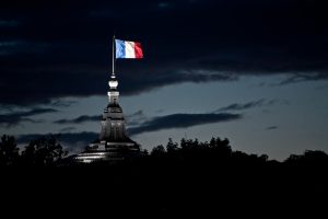 The French flag, made up of red, white and blue stripes, sits atop a tall buidling. Most of the flag's surroundings are in the shadows of the night. A light shines only on the flag.