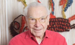 The picture shows Ted Comet, an elferly man with pale skin, white hair, and light brown glasses looks at the camera. He is wearing a red collared shirt visible to the shoulder. Behind him is a tapestry.
