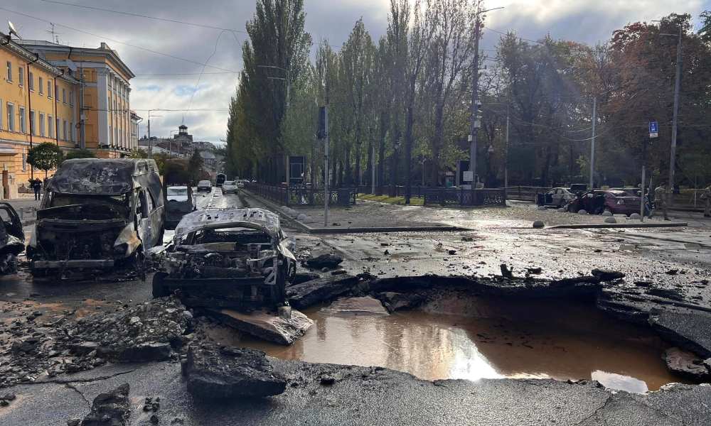 Sun breaks through clouds at a Kyiv intersection with a large hole in the pavement from a rocket blast. Destroyed vehicles sit to the left of the hole in the foreground and to the right, in the background, other cars sit empty.