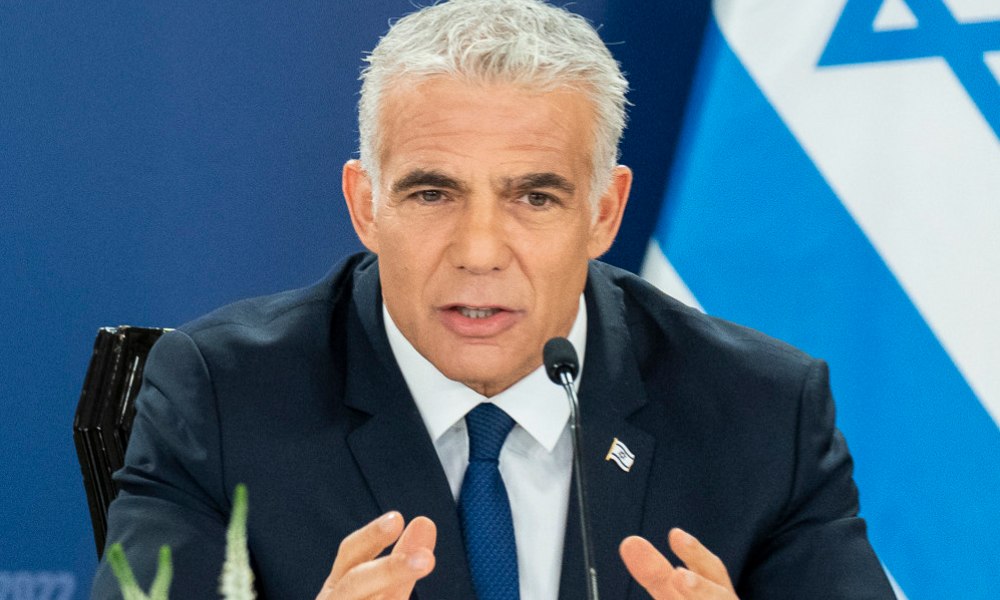Yair Lapid, a deeply handsome man with light gray hair and dark black eyebrows, speaks into a microphone while wearing a dark suit, white shirt, blue tie, and Israel lapel pin. The Israeli flag is partially visible in the background.