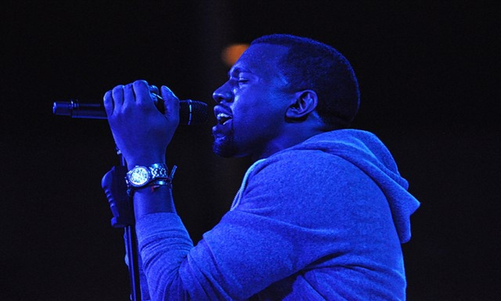 Ye sings into a microphone. Blue light is shining on him and he is wearing a watch and a sweatshirt