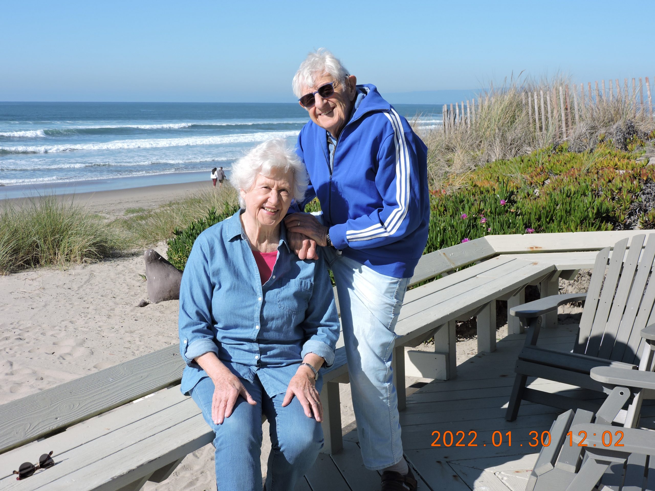 A white man with white hair and sunglasses stands behind a seated white woman with white hair wearing blue by the beach.