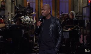 Dave Chappelle stands on the SNL stage and gives his monologue. He is wearing a black, leather jacket.