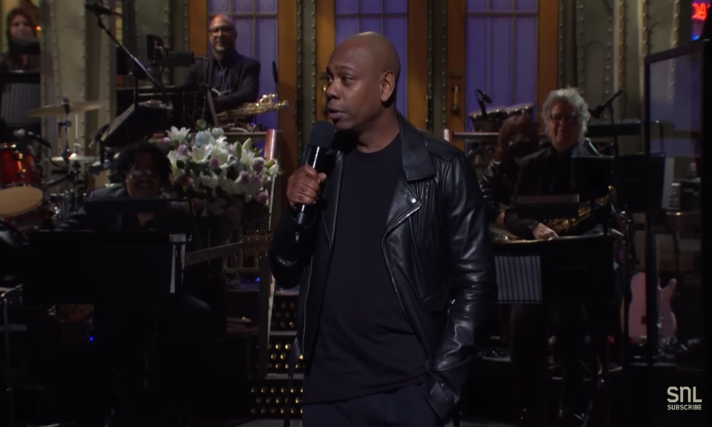 Dave Chappelle stands on the SNL stage and gives his monologue. He is wearing a black, leather jacket.