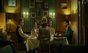 Five individuals -- three men and two women -- are sitting at a dining room table. The overall color scheme in the room is brown and green.