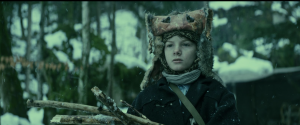 A young boy is standing in a snowy forest. He is holding some branches and wearing a fur hat and a scarf. The photo has a blueish-green glow.