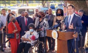 A white man in a blue suit speaks at a podium facing the camera. To the left and behind him, an elderly black woman sits in a wheelchair amid other black elders.