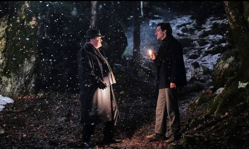 Two men stand in the woods looking at each other.