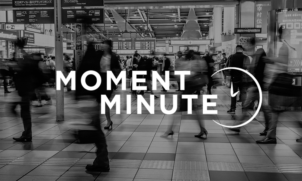 image shows a black and white blurred train station with people walking through it. The words Moment Minute and the outline of a clock overlay the train station image.