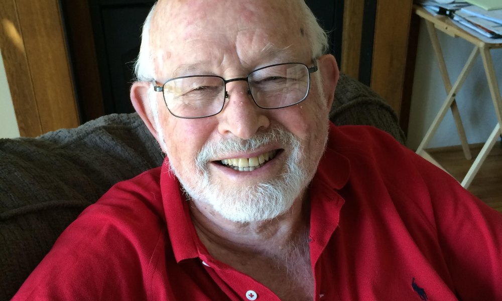Don Stone, a man with a red shirt, glasses and a closely trimmed white beardsmiles smiles at the camera