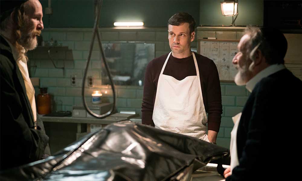 Three men standing in a funeral home with a body bag on a table in front of them