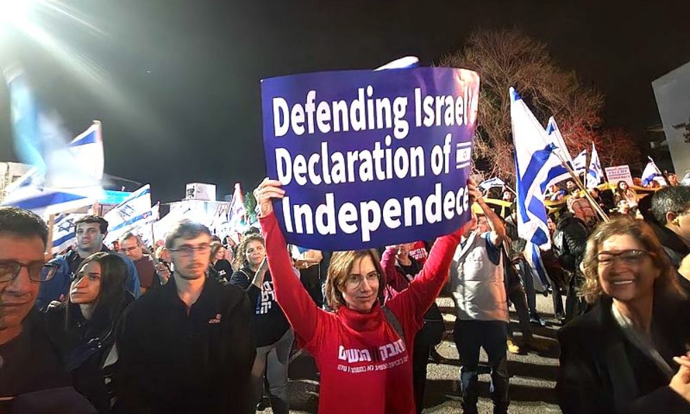 A woman in a red sweatshirt holds up a blue sign which says "Defending Israel's Declartion of Indepence" at a protest in Haifa.