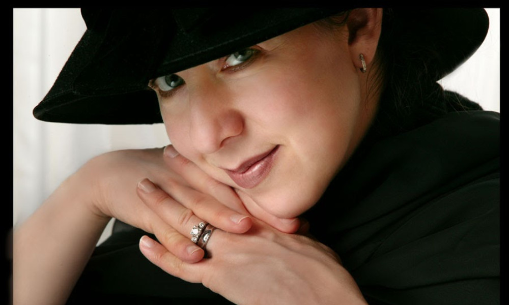 A headshot of a woman resting her face on her interlocked fingers. She is wearing black clothes and a black hat, and she wear a wedding and engagement ring.