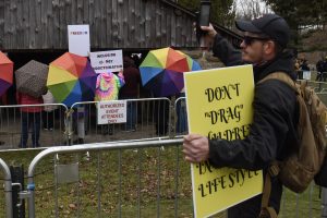 A man in dark clothing lifts his phone to videotape a group of people behind a barricade, who are holding rainbow umbrellas. The man holds a yellow sign which says, in part, "Don't 'drag' children into [your] lifestyle."
