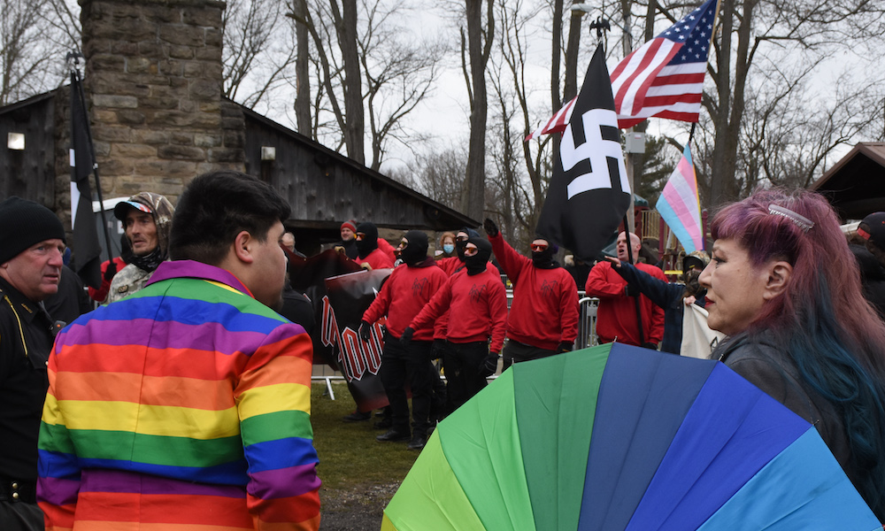 A man in a rainbow jacket looks at a crowd of men wearing matching red sweatshirts with black ski masks and pants. One makes a Nazi salute, another holds a swastika flag and American flag.