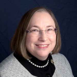A headshot of a smiling white woman with light brown hair, a pearl necklace, and a dark blue background.