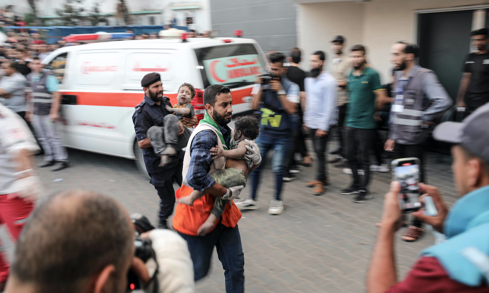 Two medics carry children into a hospital surrounded by onlookers.