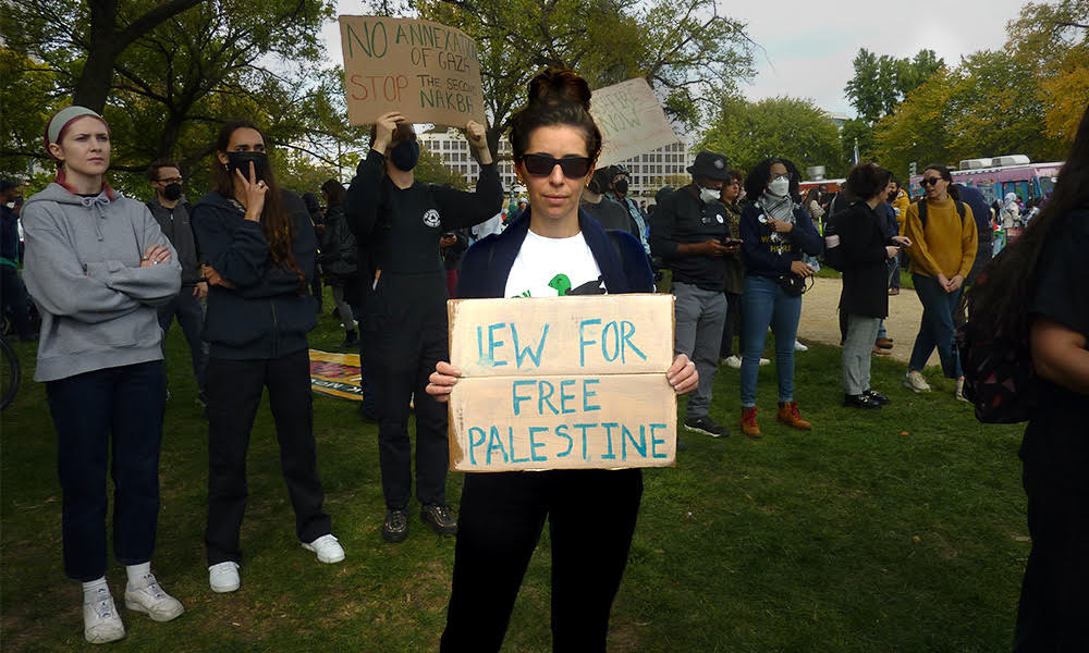 A woman wears sunglasses and holds a sign that reads JEW FOR FREE PALESTINE written in blue ink on cardboard.