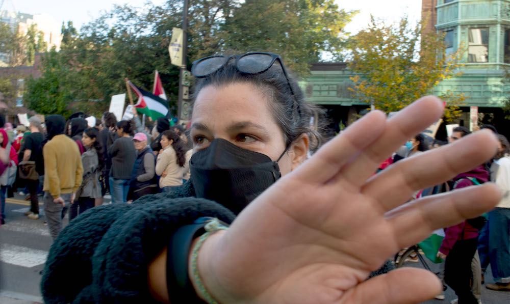 A masked white woman holds her hand up in front of the camera lens. On the street behind her, doxens of people march, some with Palestinian flags.