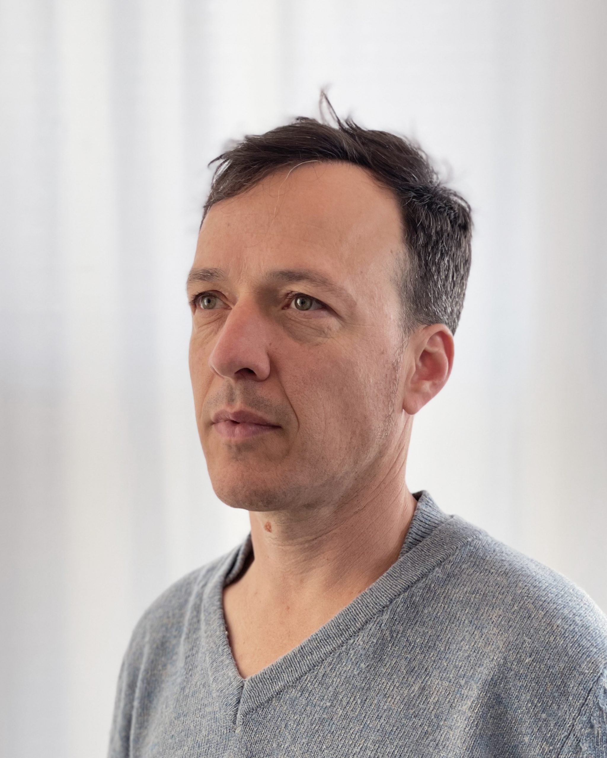 A white man with short dark hair wearing a gray v-neck t-shirt gazes to the left of the frame