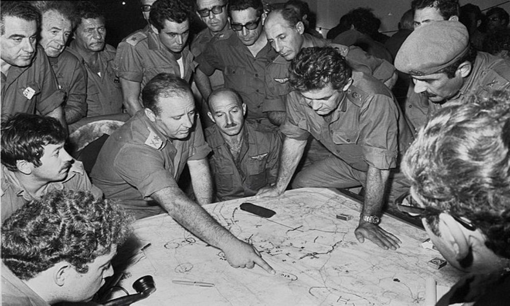 A black and white image of a group of men looking at a map. One man is pointing to a specific point on the map.