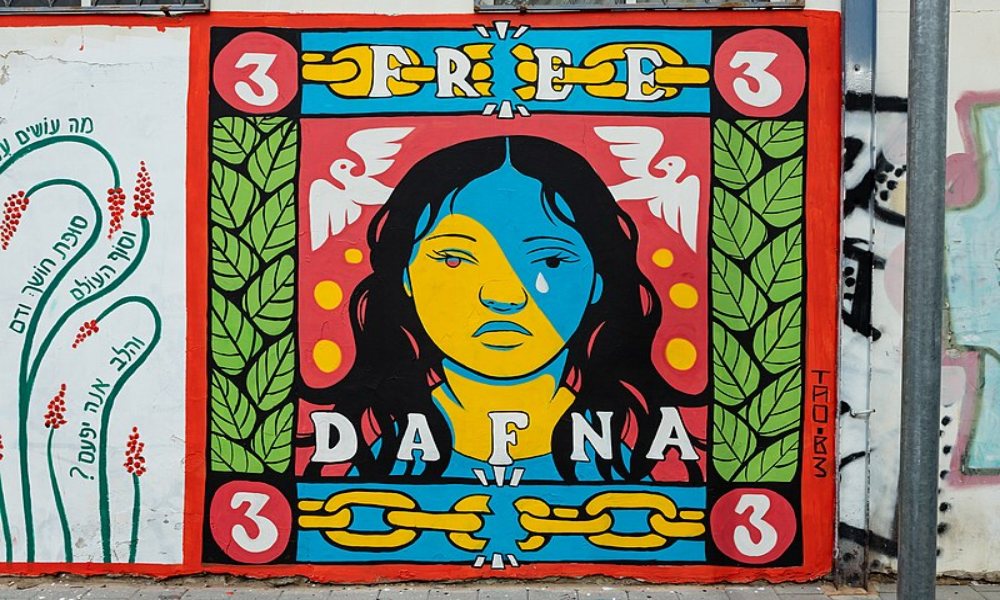 A colorful mural on a wall depicts a girl's face, half blue and half yellow, surrounded by leaves and doves. Above her head is the word Free on top of a chain, and below her head is the name Dafna, also atop a chain. The letter 3 is painted in each corner of the square mural.