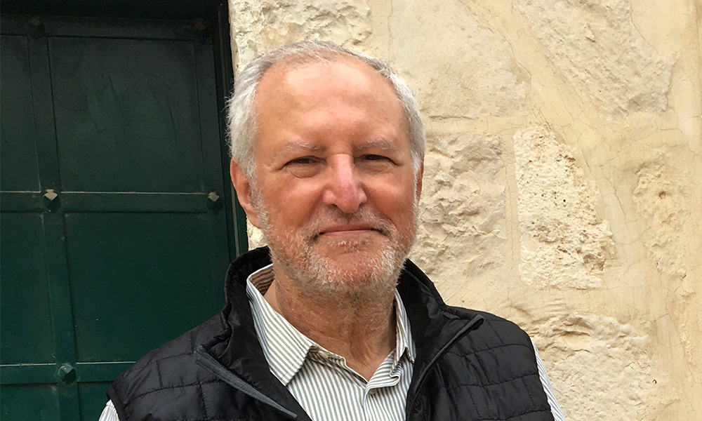 Mohammed Dajani, an olive skinned man with white hair and stubble wearing a collared shirt and a black down vest, smiles at the camera in front of a stone building with a green door.