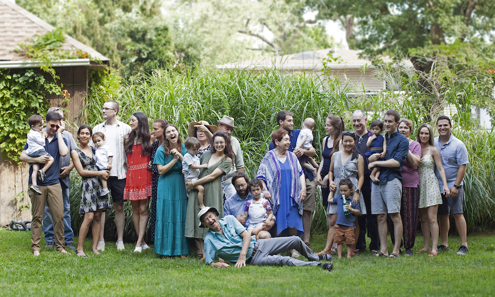 A tall elderly man sprawls happily on the ground in front of a large intergenerational crowd of family members in front of some green foliage.