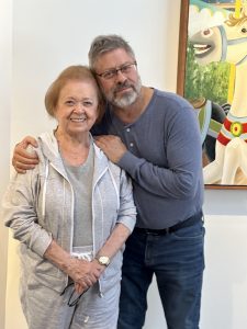 An older woman in gray smiles. A bearded man hugs her warmly. They are standing in front of a painting.