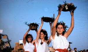 Young women wearing white gowns and flower crowns hold baskets of greenery over their heads.