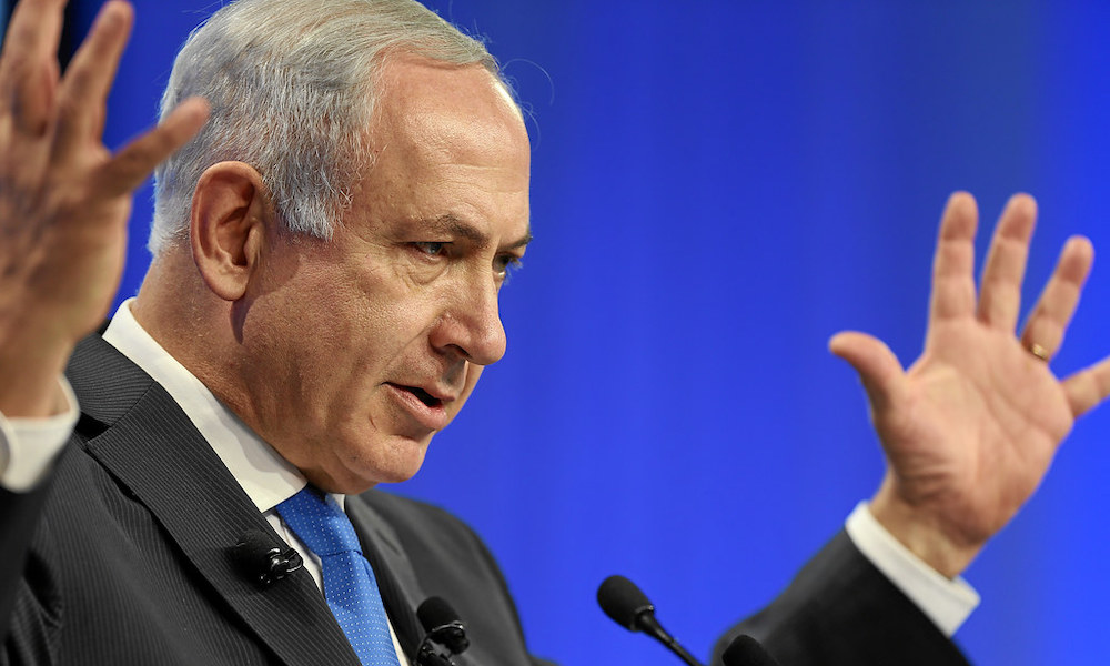 Israeli Prime Minister Benjamin Netanyahu in a suit with a blue tie, speaking with hands outstretched.