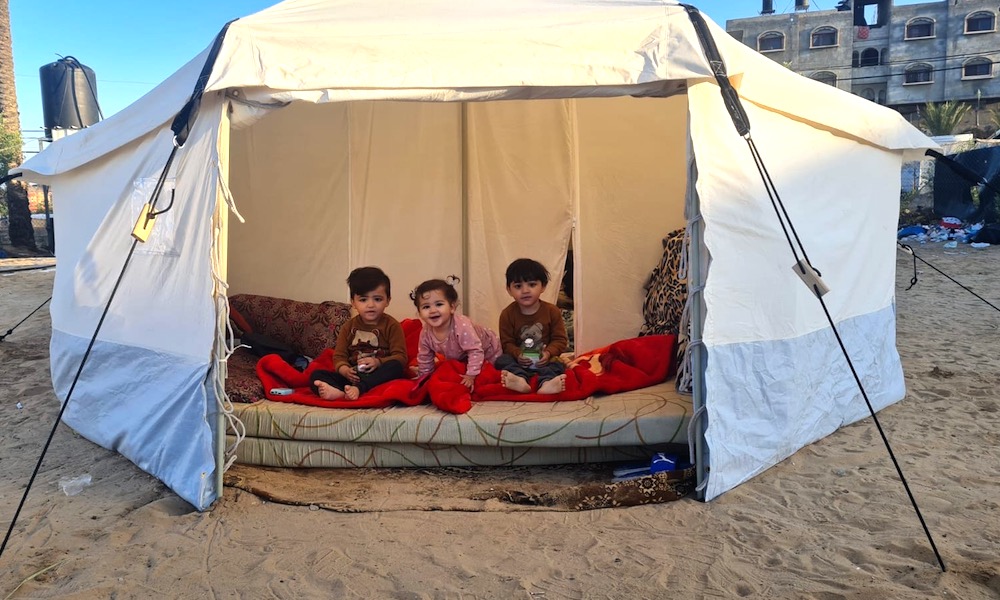 Three small children sit in a tent on a beach.