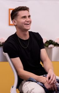 Young man in a black T-shirt sits and smiles.