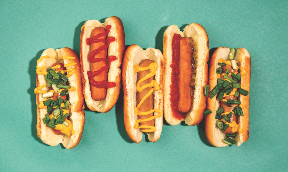 An illustrative photograph depicting five colorfully topped hot dogs in buns on a turquoise background.