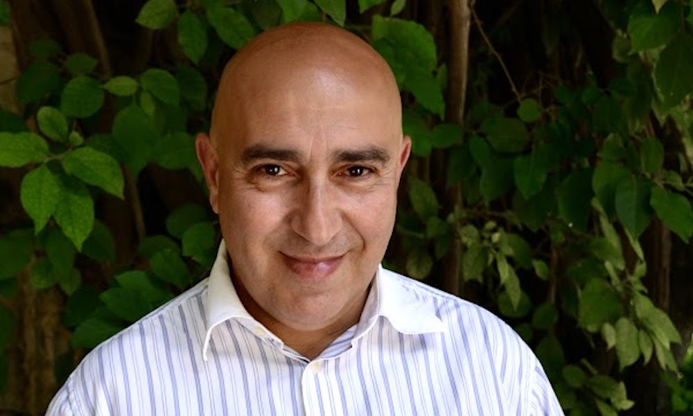 A smiling Palestinian man with a clean-shaved head and dark eyebrows smiles at the camera wearing a crisp button-down shirt. In the background is foliage.