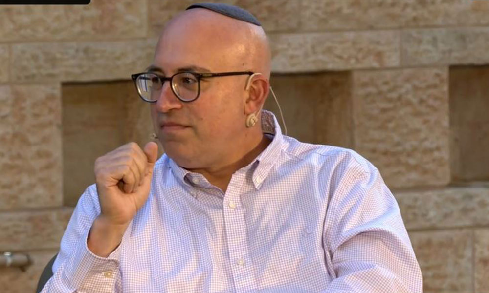 A bald man with a black yarmulke and a pink button down shirt with black rimmed glasses looks to his right and holds his hand over his face